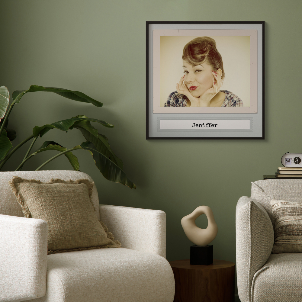 Vintage portretposter met naam in een zwarte lijst aan een groene muur naast een plant en lichte fauteuil
