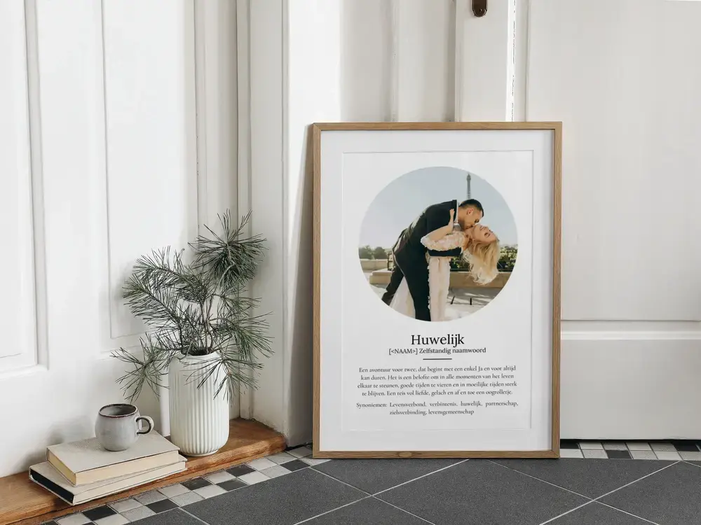 Poster in woordenboekstijl over het huwelijk met ronde trouwfoto, in houten lijst op de vloer naast een plant