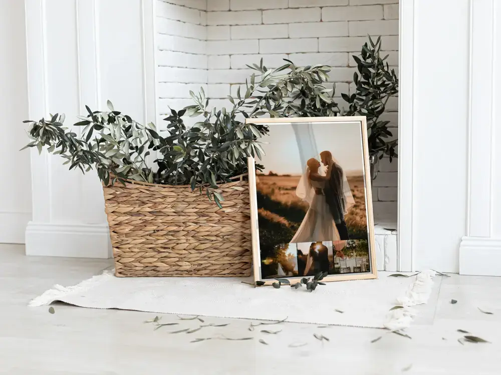 Staande trouwfotocollage in een lichte lijst, op de vloer naast een plantenmand voor een witte bakstenen muur