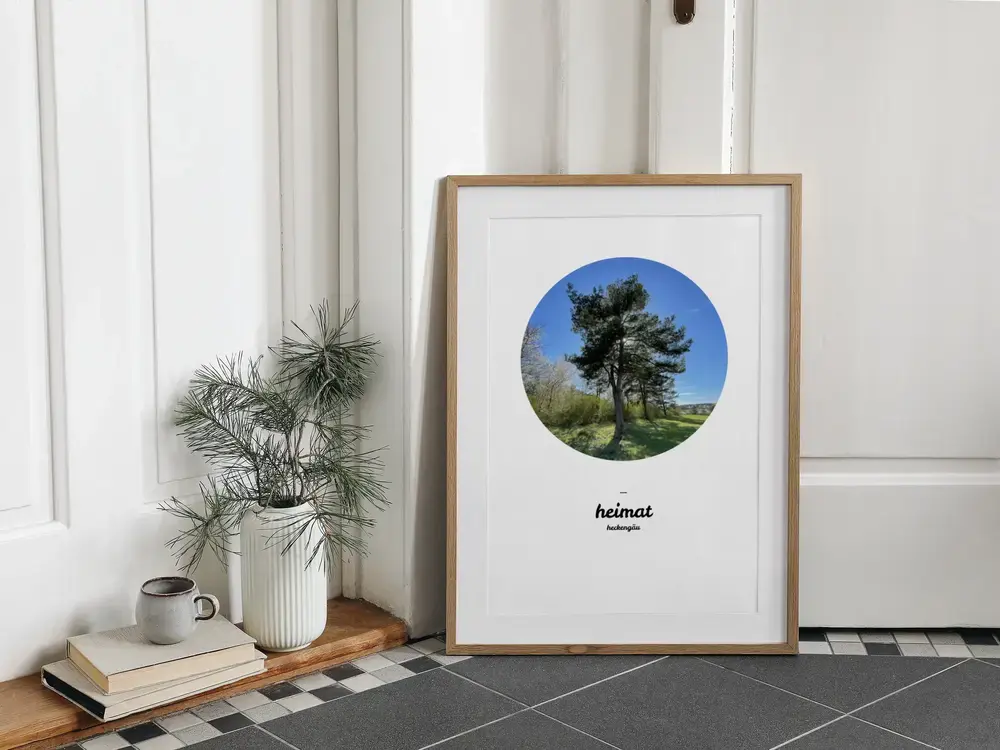 Heimat-Poster mit rundem Kiefernmotiv und individuellem Ortsnamen im hellen Holzrahmen, am Boden neben einer Vase