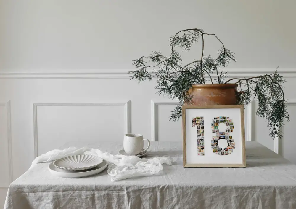 Pêle-mêle photo en forme de 18 dans un cadre en bois, sur une table dressée près d’un vase de branches