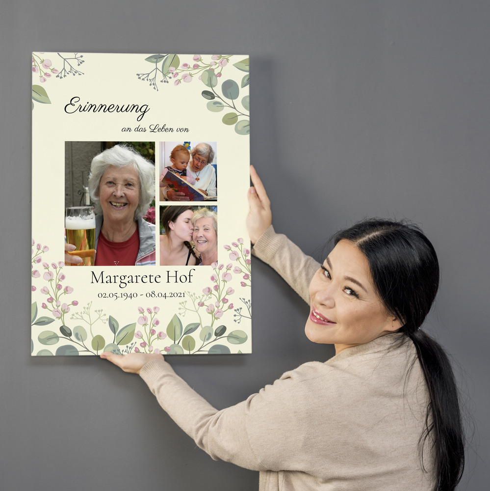 Personalisierte Trauercollage auf Leinwand mit drei Fotos und zartem Blumenrand vor grauer Wand